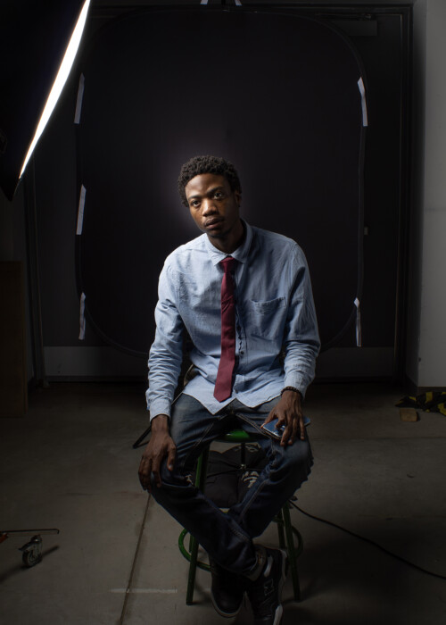 Subject named Osi wearing a blue collared shirt with a bergundy tie, sitting on a green stool with while a light is shining in front of him. Behind him is a door that has the label 'exit' above it.
