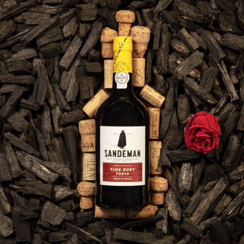 A wine bottle with a red label that reds 'fine ruby porto' laying on a bed of corks. Black wood chips surround the bottle, and a red rose lays on the right side of the bottle.