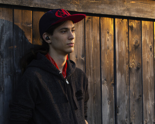 A person is leanng against a black fence, hands in their pockets and listening to music with their earbuds on as the setting sun shines down on them.