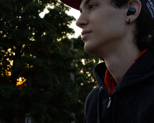 Person looks out into the distance longingly as they listen to music. The orange sun beside them glistens through a trees leaves and branches.